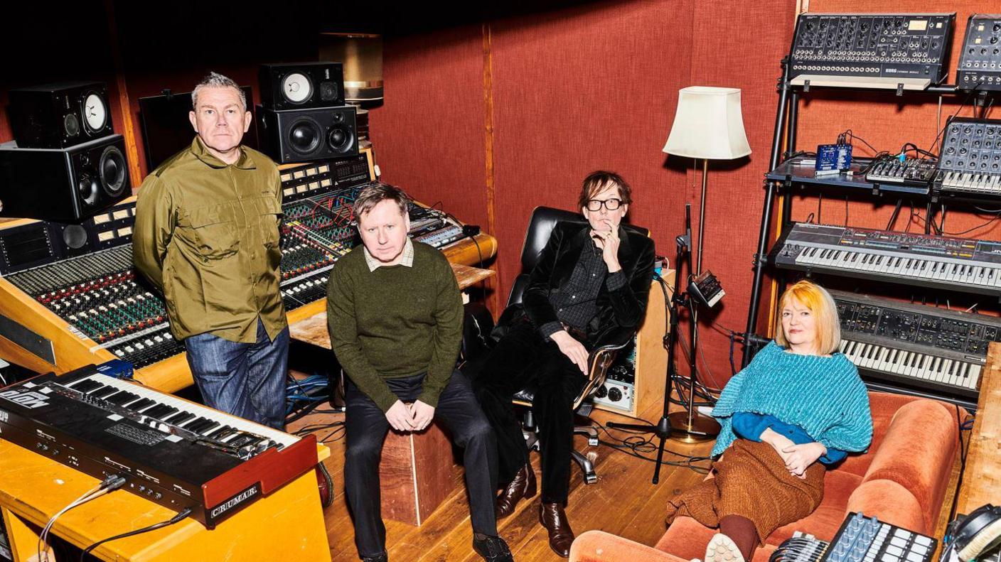 Jarvis Cocker sits in a chair with two other male members of Pulp to the right of him, standing up, and Candida Doyle is on an orange velvet studio to the left. All are in a music studio.

