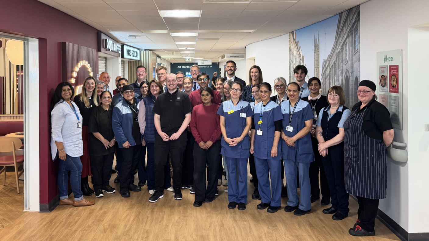 A large group of people inside a new hospital entrance. One wall on the left is red-maroon and has a costa coffee and M&S food signs.