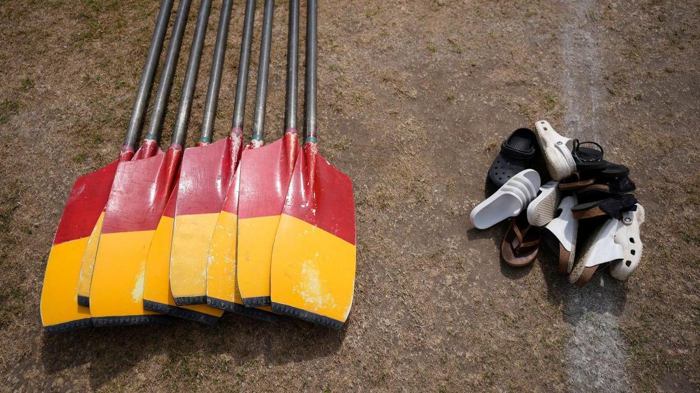 A collection of black, red and yellow oars laid on some grass on the left hand side of the frame, with a pile of shoes on the right.