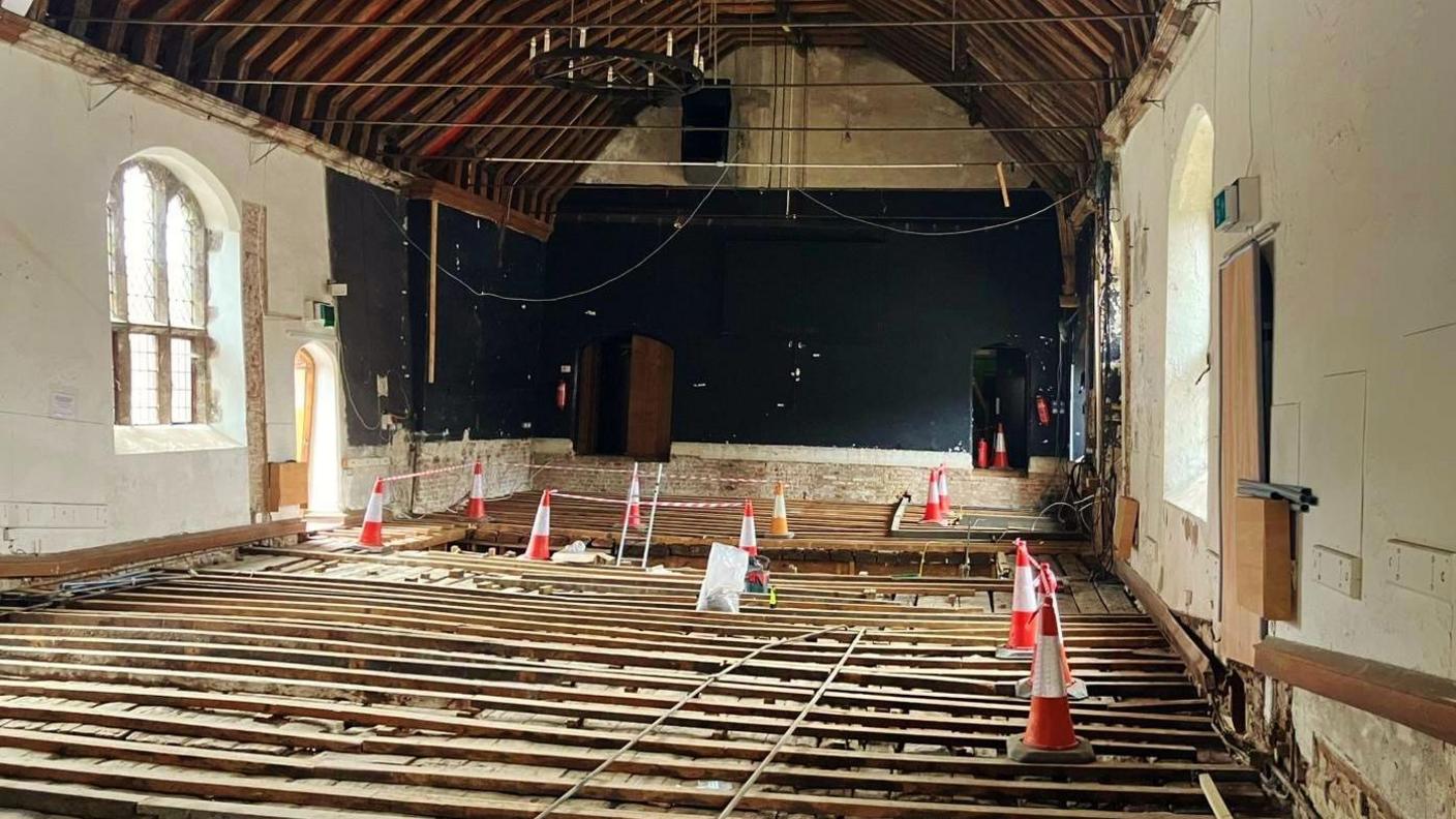 Looking towards the back of the theatre where there are cones on the floor boards. Two doors are at the end of the building, and the ceiling is arched with wooden beams.