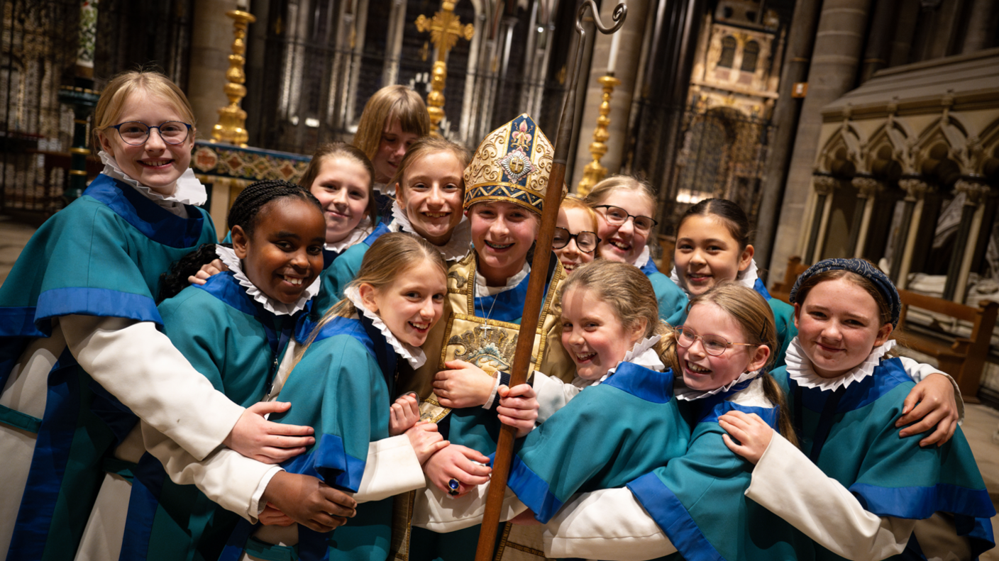 A dozen girls in the cathedral wearing chorister uniform of white undercoats with blue tunics on top, all smiling to camera and hugging Emmie Piper who is wearing vestments similar to a bishop and a mitre