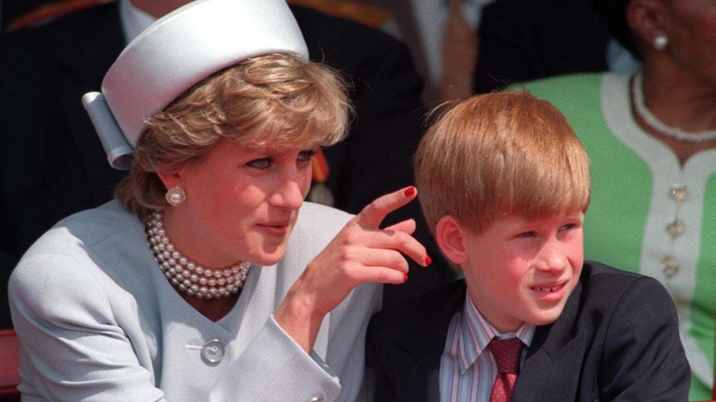 A young Prince Harry is pictured with his late mother, Diana, Princess of Wales, in 1995.  She is dressed in a pale jacket and matching hat. She has short hair and is wearing pearl earrings and a necklace. She is pointing to show something to Harry who is looking into the distance. He is wearing a dark suit jacket, a striped shirt and a red tie.