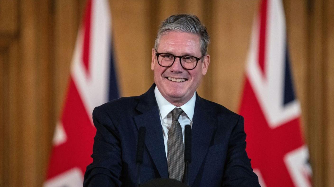 Sir Keir Starmer standing in the Number 10 briefing room. He is smiling at the crowd. He wears a dark suit, white shirt, grey tie and black-rimmed glasses. Behind him are two union jack flags.