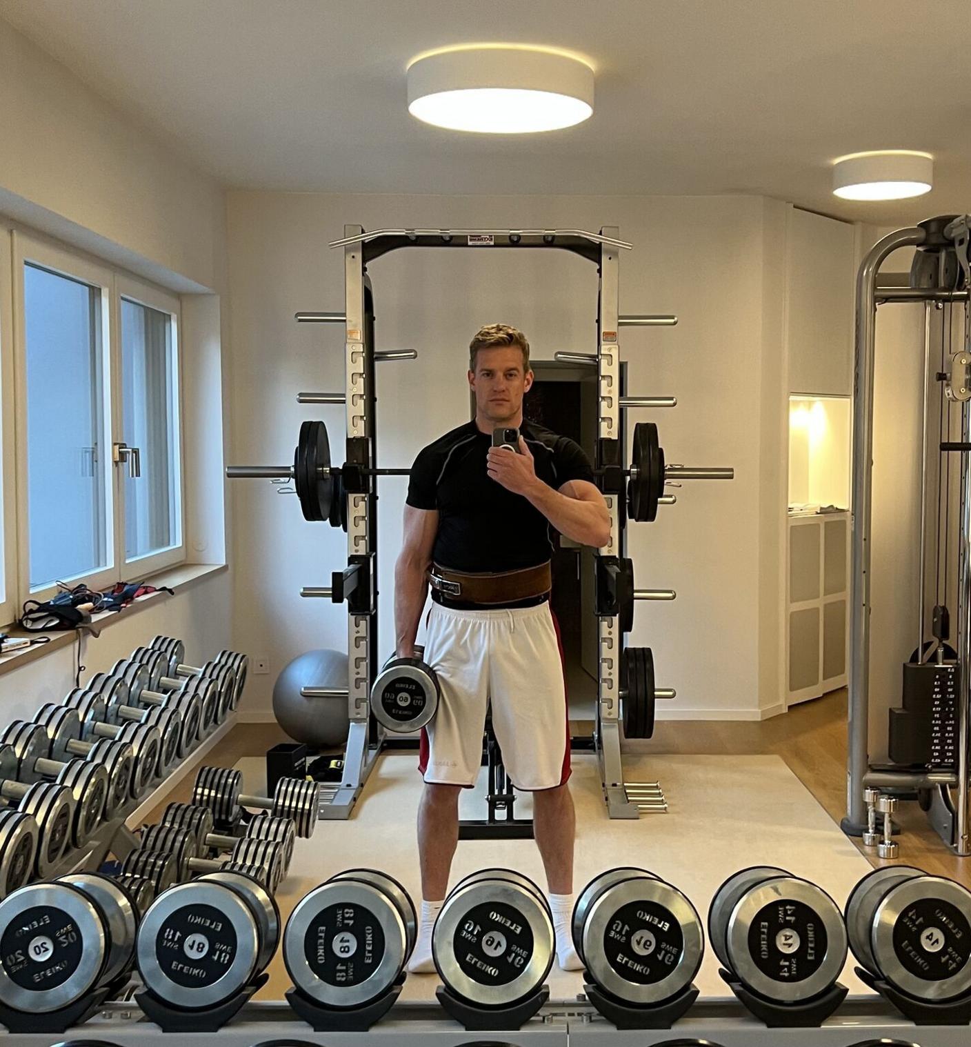 Stefan Hoops takes a selfie of himself holding a weight in one hand and surrounded by heavy weights in a gym