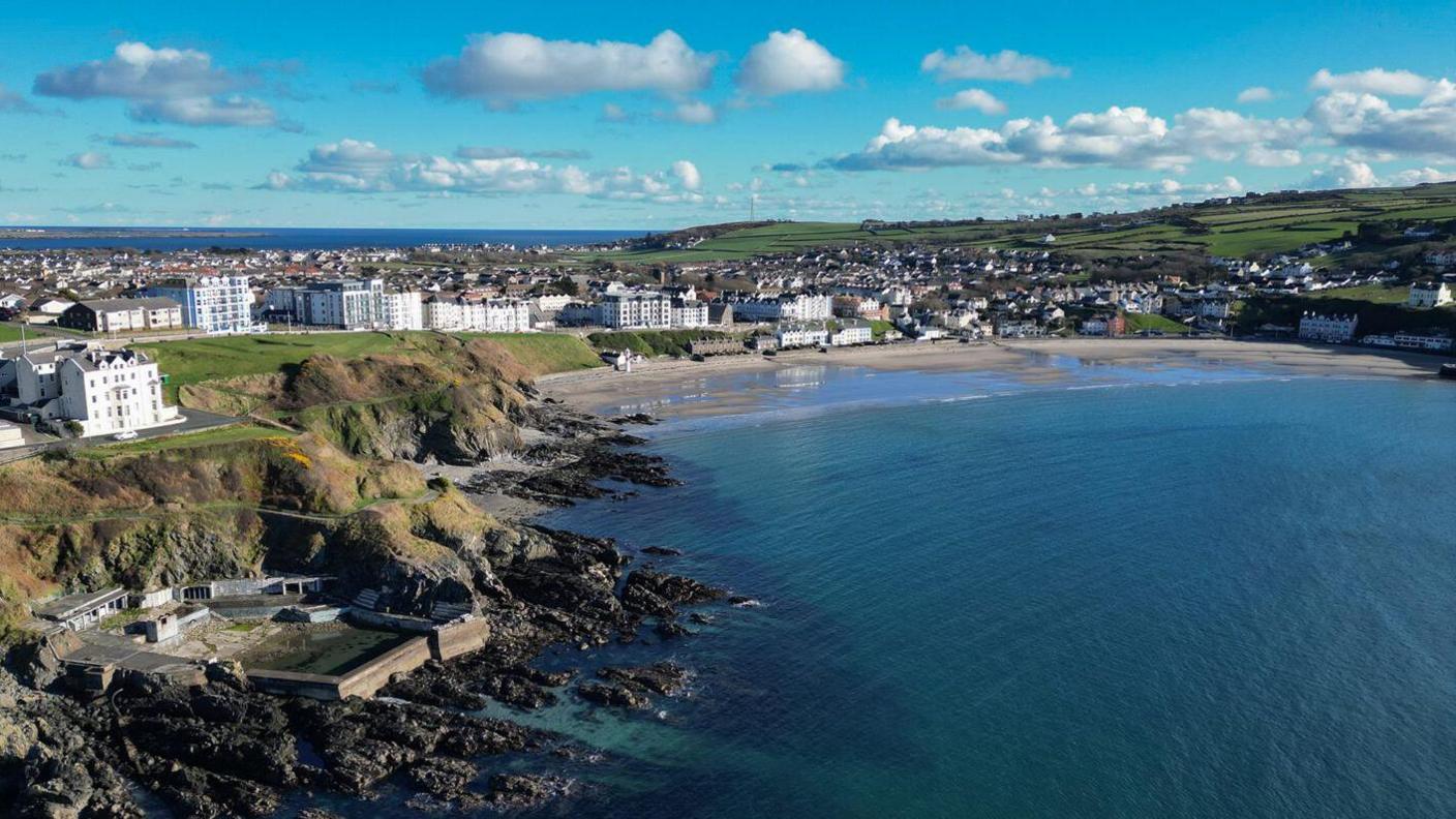 An aerial view of Port Erin. Rows of tall white buildings line the seafront with green hills behind them with Port Erin Bay below. It has a sandy beach which meets a turquoise-coloured sea. The sky is blue with white fluffy clouds. 