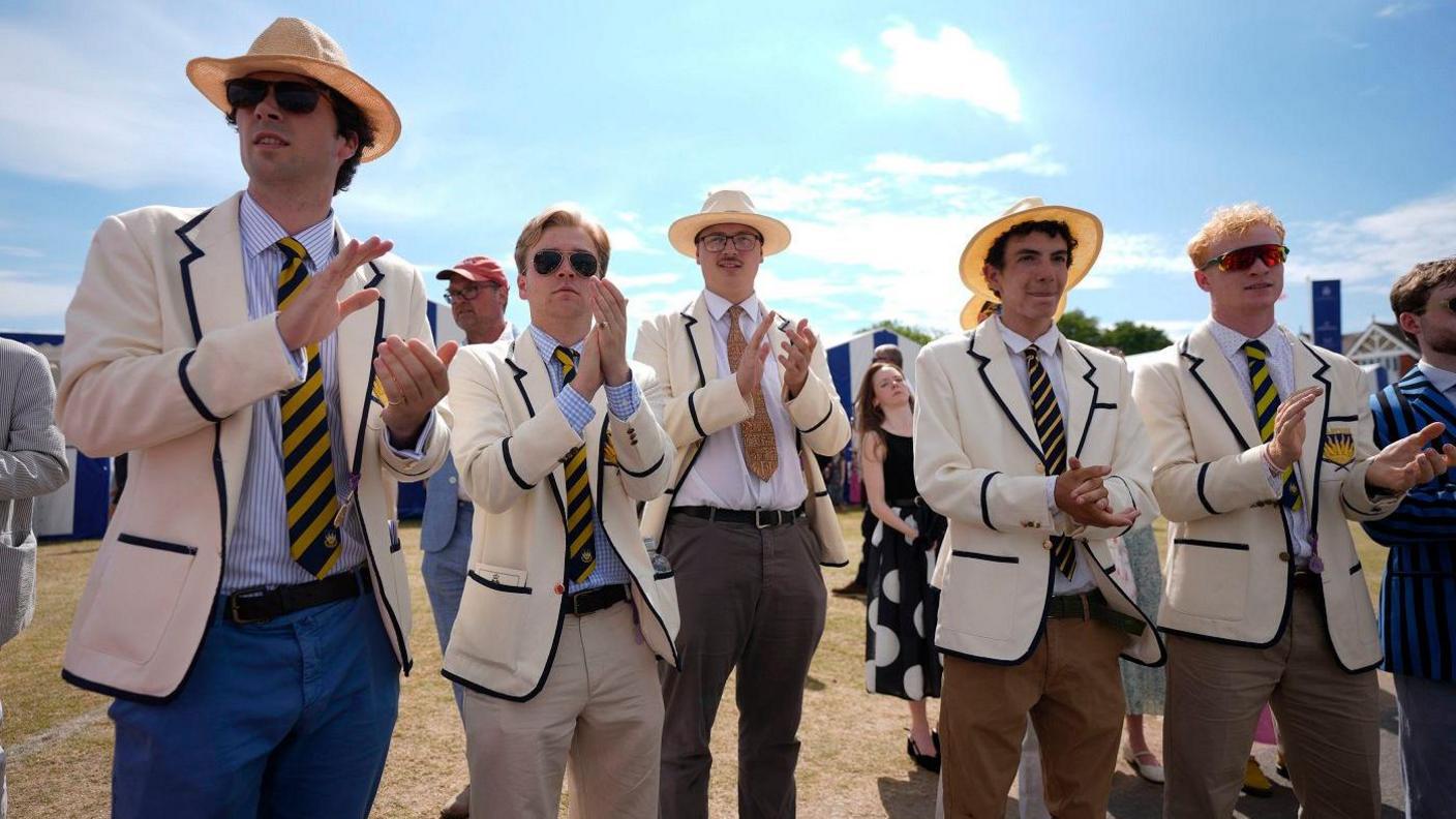 Five men in suits, white blazers and wicker hats smiling and applauding something off camera. It's a sunny day and the sky is blue.