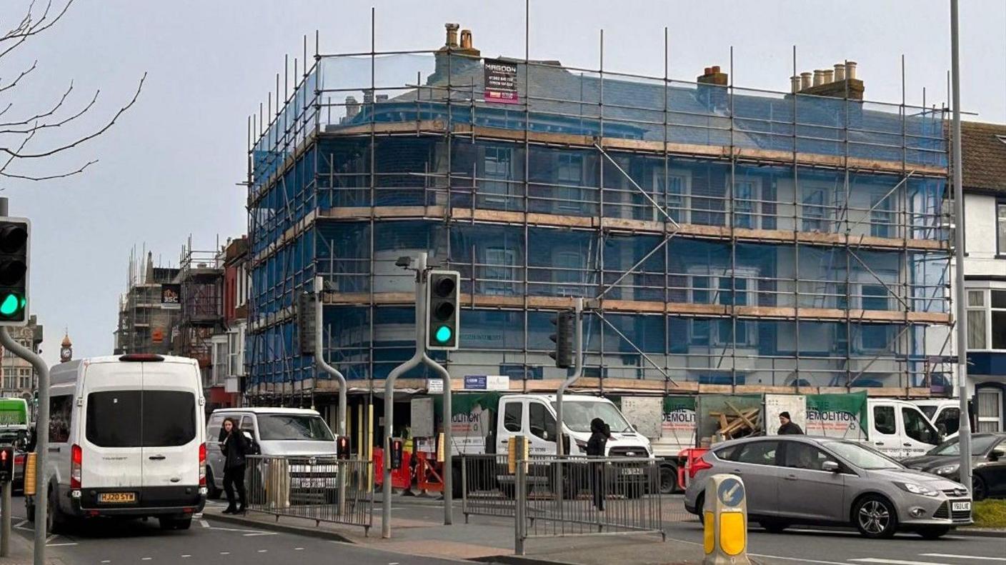 Scaffolding covers the former pub building - a three storey Victorian corner terrace with a pitched roof. The road in front is busy and filled with cars and vans.