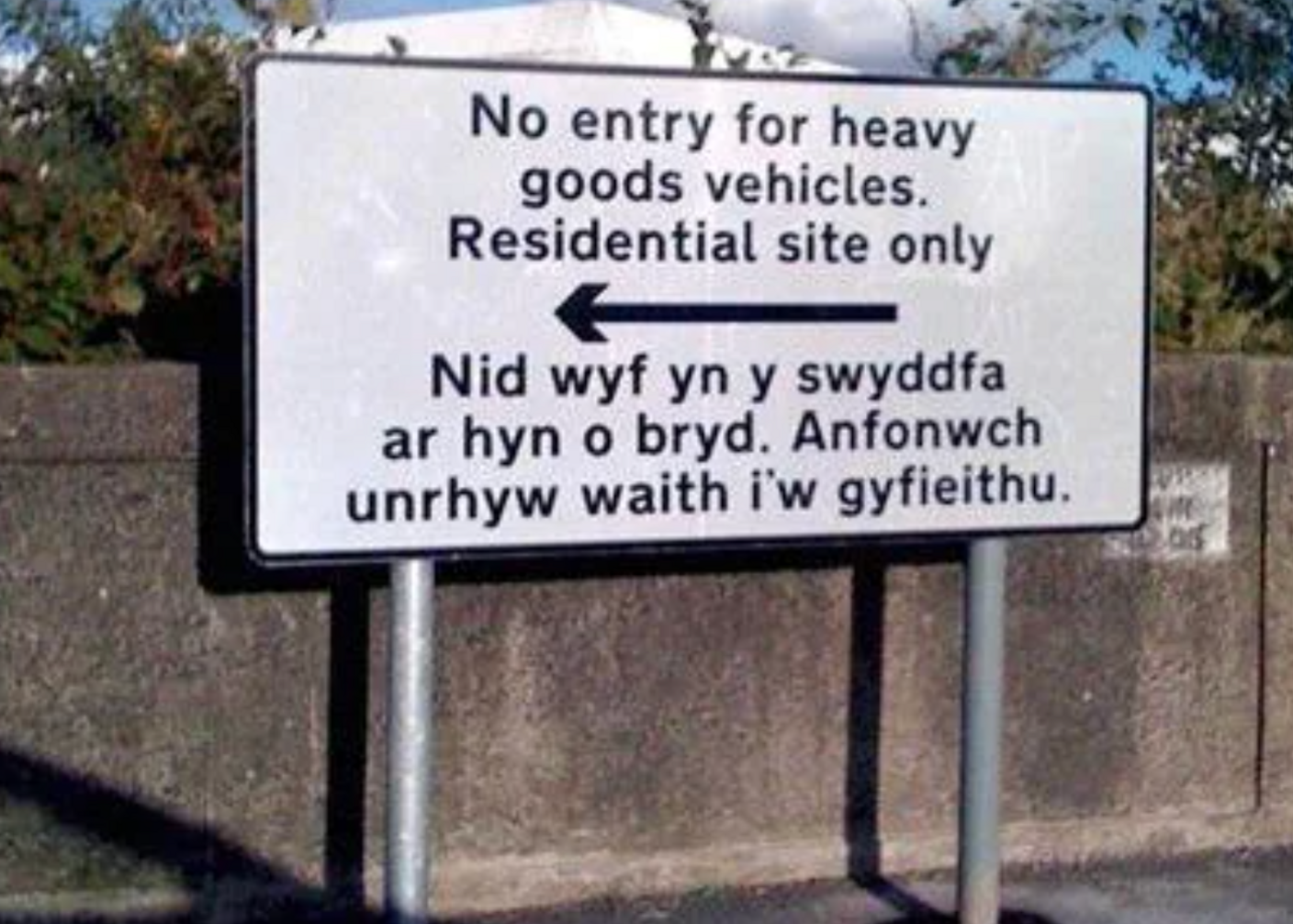 A white sign with black lettering that reads: No entry for heavy good vehicles. Residential site only, followed by Welsh that reads: "I am not in the office at the moment. Send any work to be translated."
