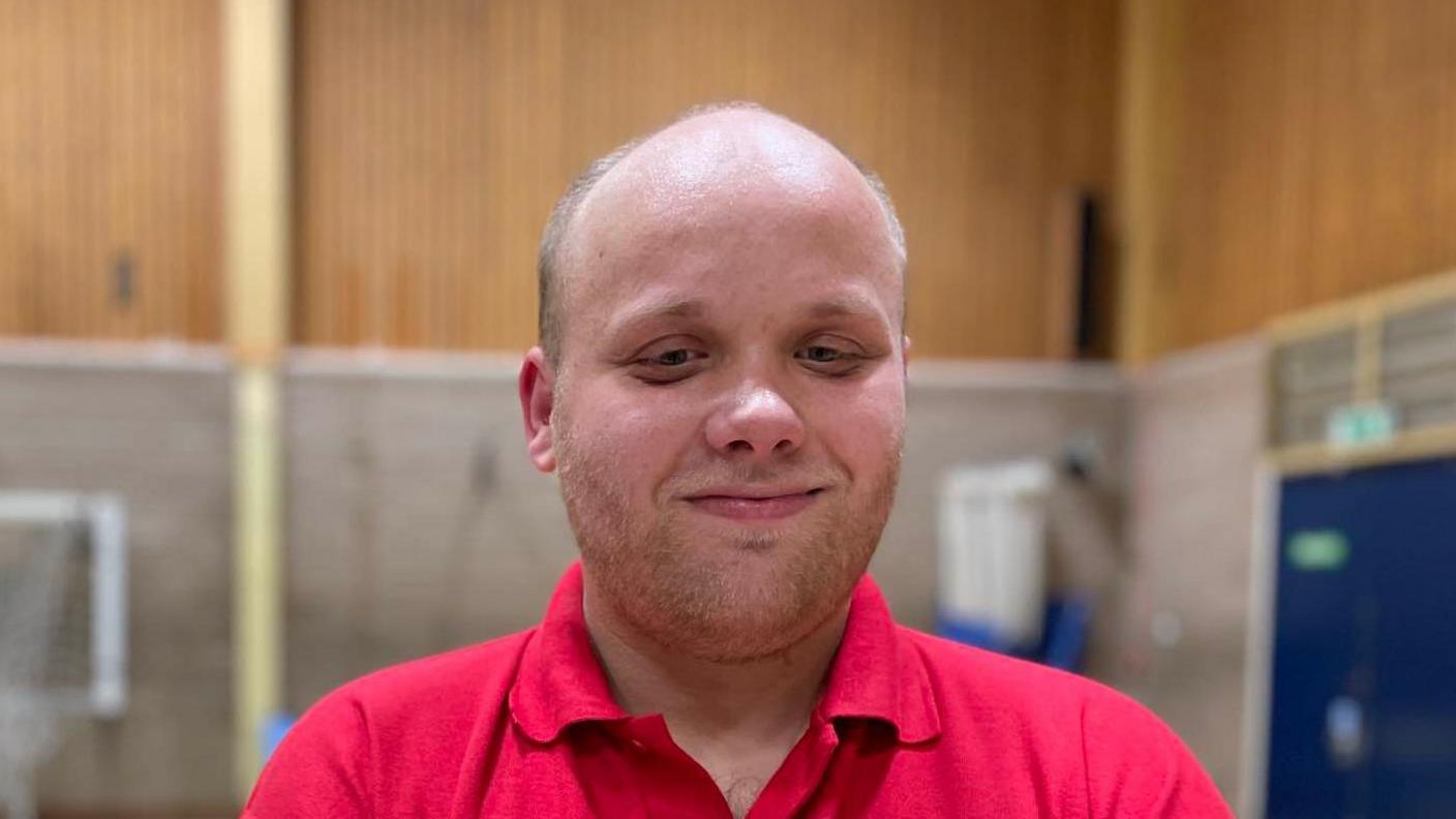 Matthew Foulger smiles at the camera while standing in a sports hall. He is wearing a red polo top.