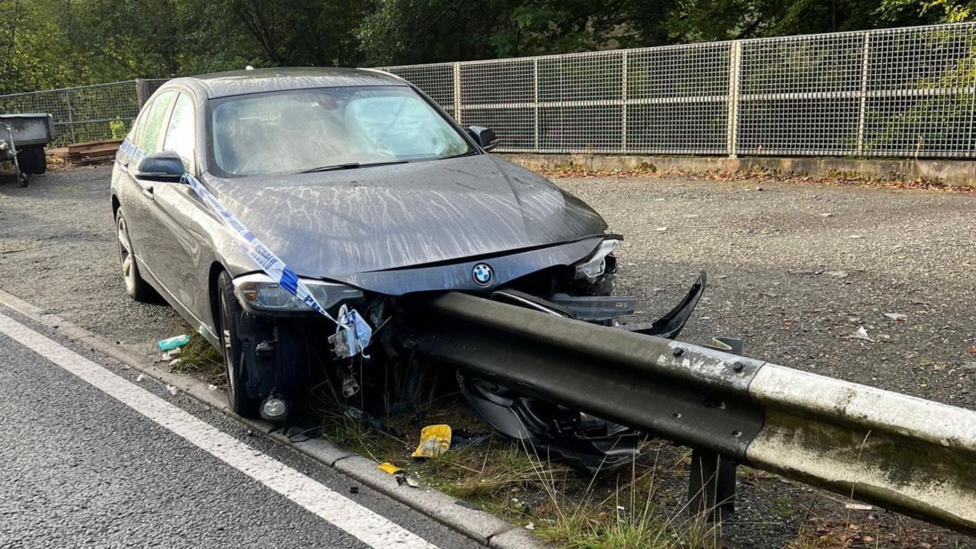 A black car has collided into a metal guardrail that was on the side of the road. Parts of the front has been significantly damaged with the bumper coming off. There is blue police tape on the side of the car. 