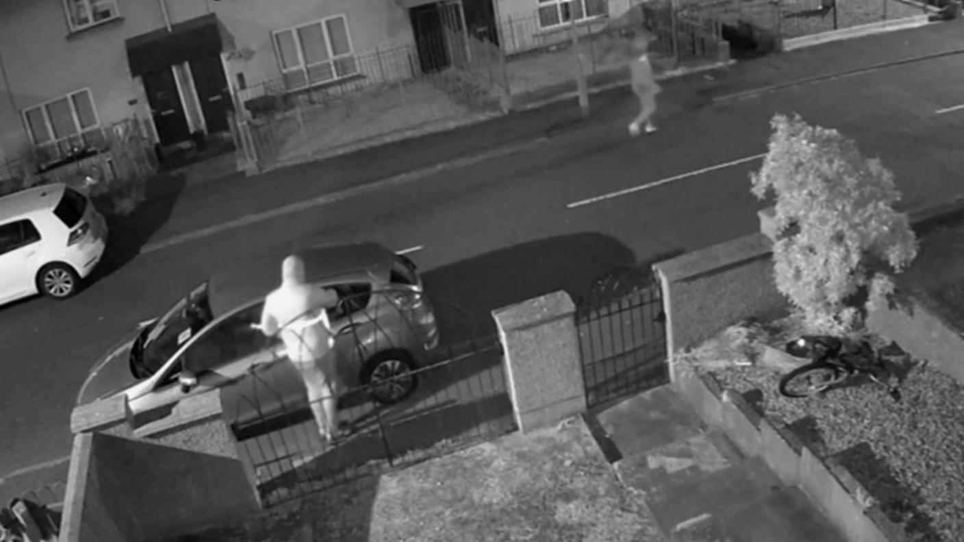 Black and white CCTV still image showing a man attacking a car wearing a light hooded jacket. Another assailant can be seen further up the street