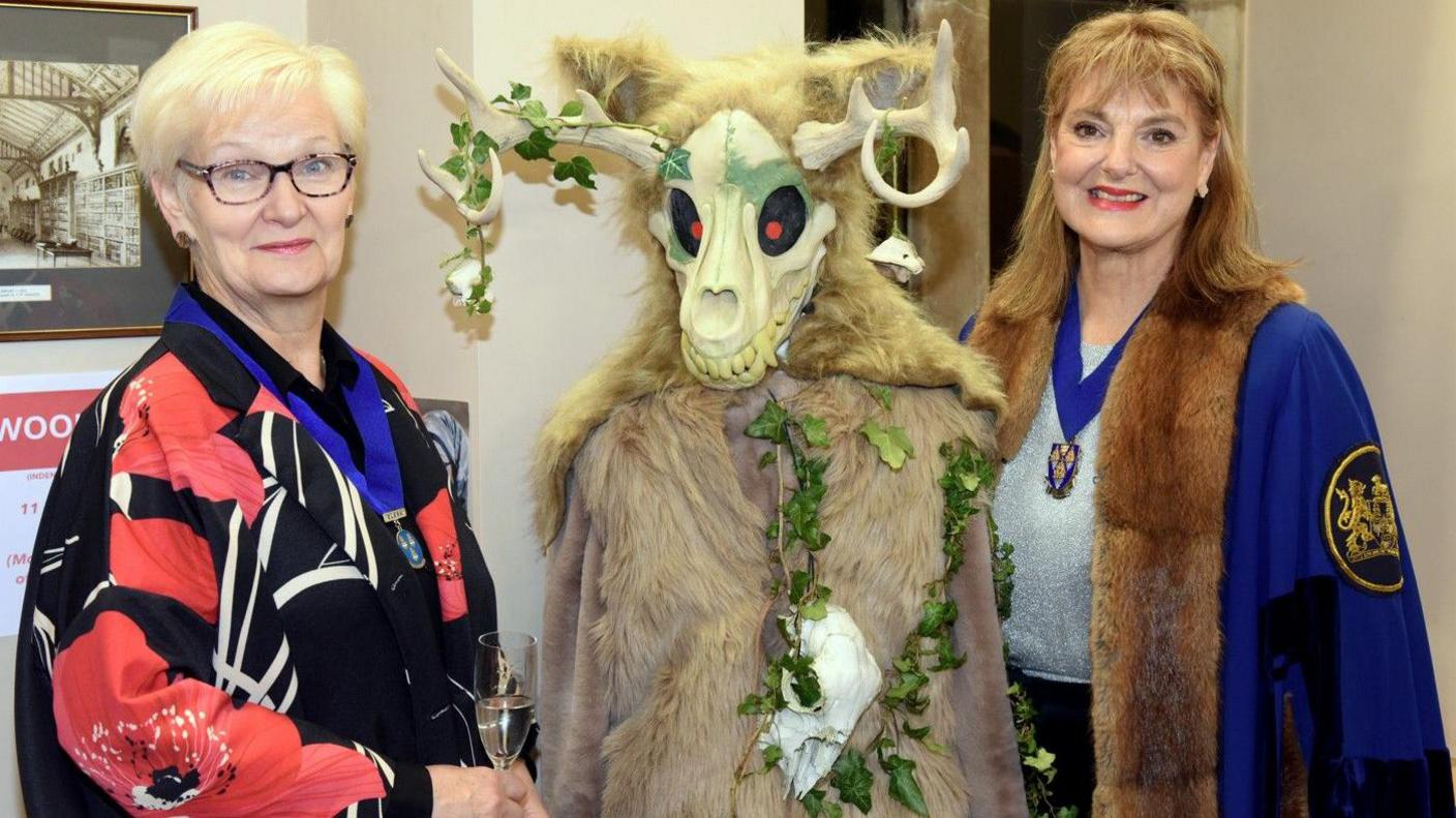 Shrewsbury Drapers Company clerk Lyndsey O'Loughlin standing on the left with short grey hair and wearing a medal on a thick blue ribbon. Master draper Nancy Sykes-Waller standing on the right wearing a blue fur-trimmed cape and medal. Between them is a mannequin wearing a fur costume and skull face with ivy leaves wrapped around it created as one of the competition entries