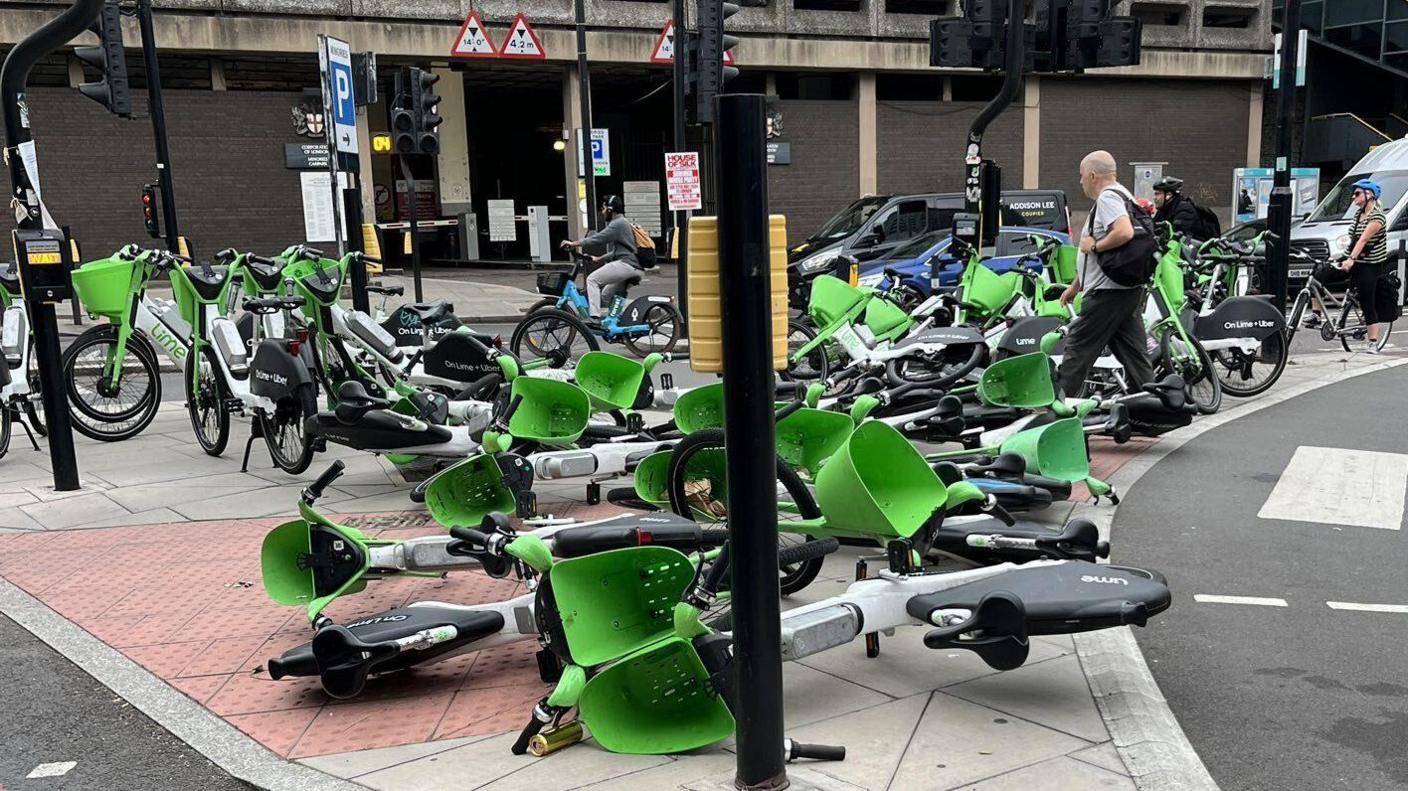 Around a dozen dockless e-bikes lie on their sides on a traffic island in Tower Hill where several more have been crammed into a small area of pavement that is blocking pedestrians from crossing