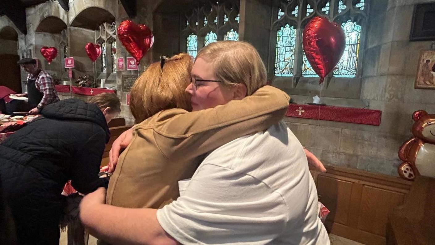 Natalie Walsh and Jackie Southall are hugging each other. They are in a church with lots of red balloons tied to the windows.   