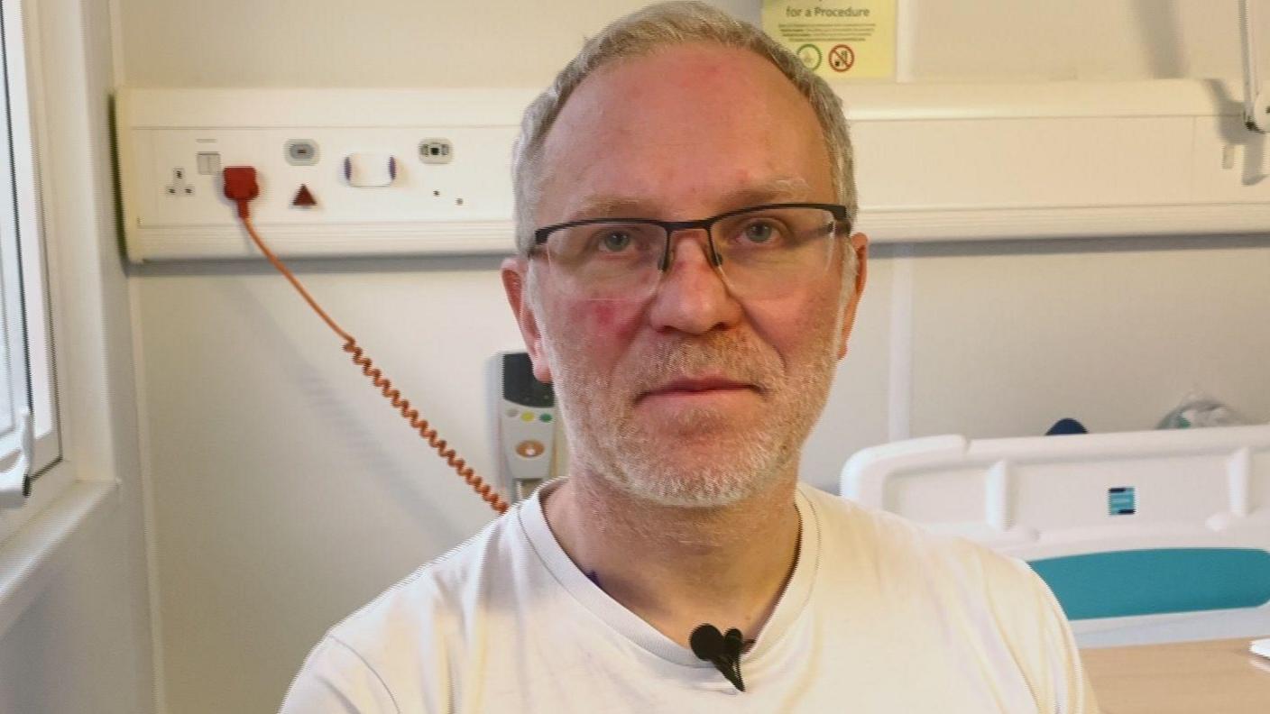 A man in a white T shirt wearing glasses sitting in a hospital ward. 