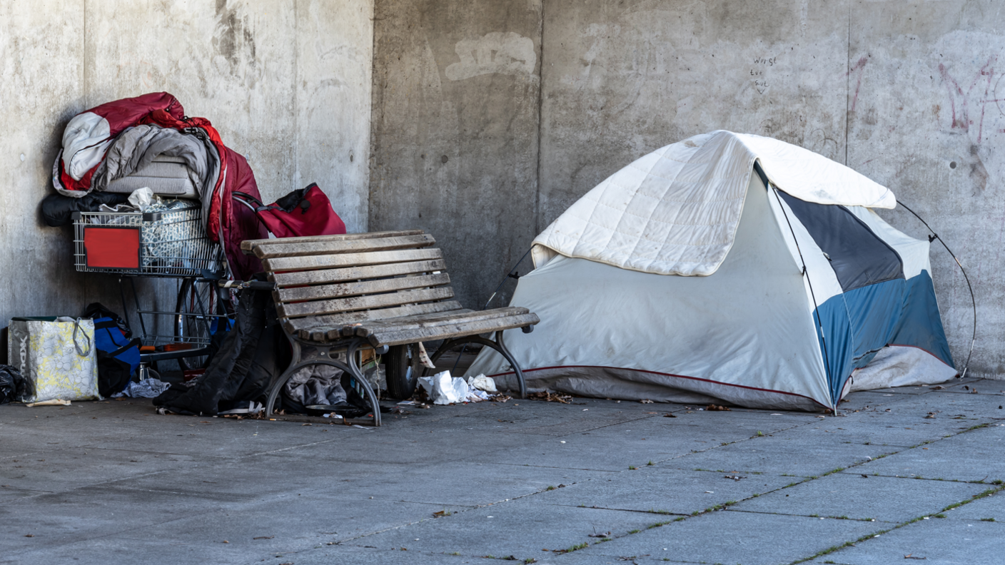 A tent and the belongings of a homeless person next to a bench outside