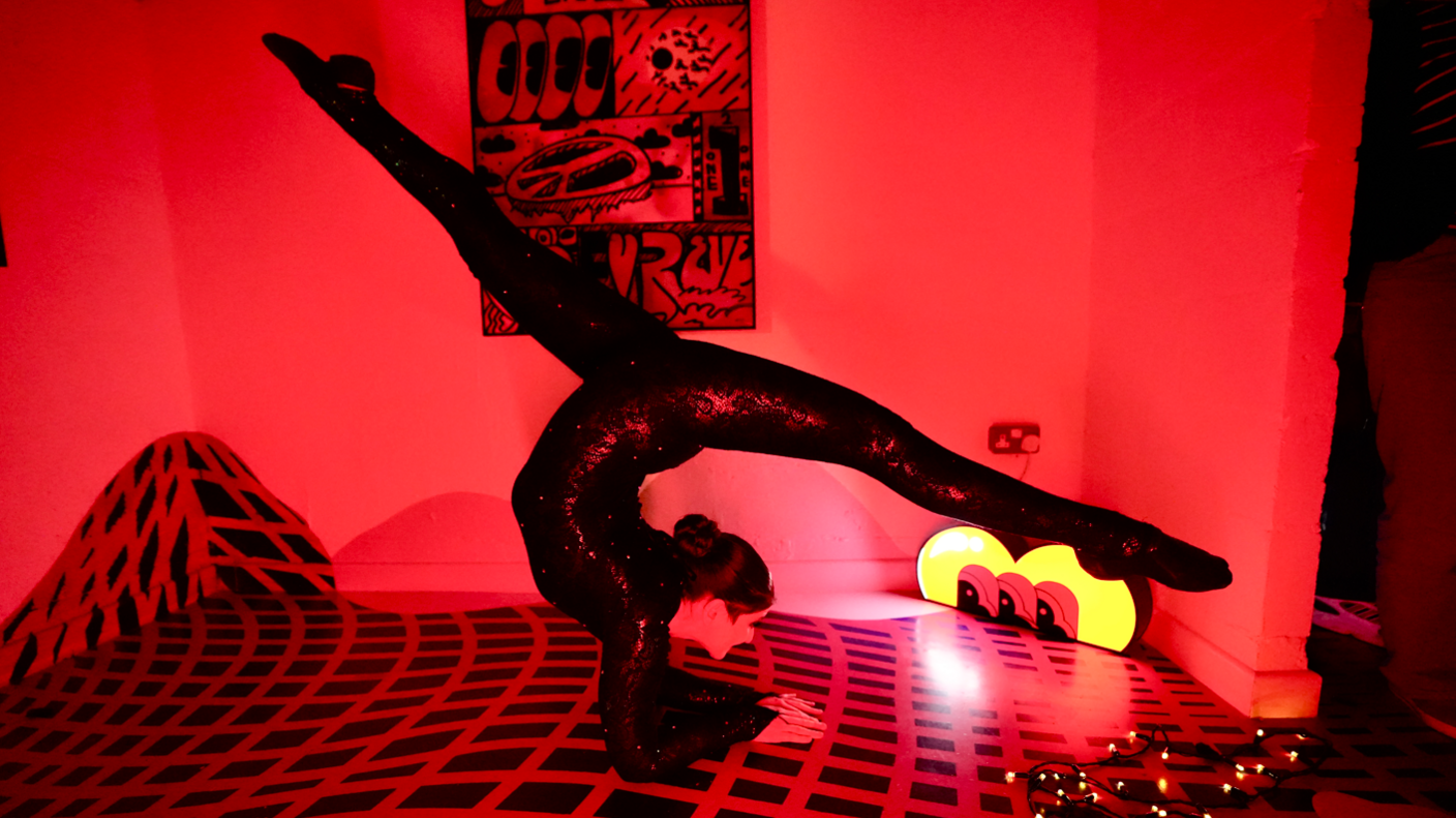 A contortionist in a spangled leotard with her forearms on the floor and doing the splits in the air in a room with red lighting and a distinctive red and black chequered floor