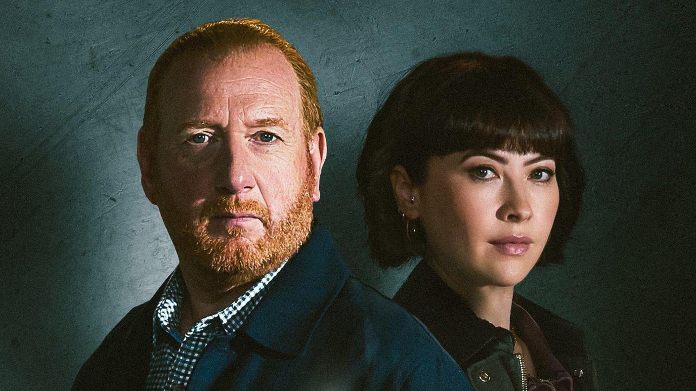 A promotional shot of the TV show with Adrian and Vanessa both looking at the camera. Adrian has ginger hair and a beard and moustache, and wears a blue overcoat and checked blue shirt. Vanessa has short thick black hair and wears earrings, and a black coat