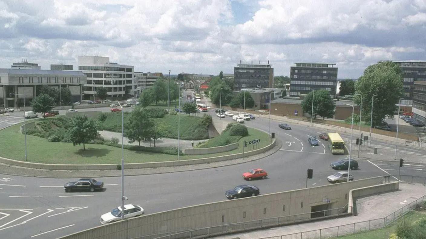 A view from a high position of the roundabout with cars on the road around it and office buildings around the edge of the roads. In the middle is a grassy area with small trees.