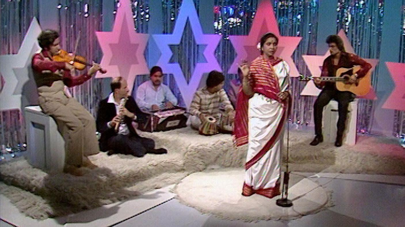 A pistillate successful a achromatic and reddish sari singing into a microphone portion musicians are sat down her playing instruments. The signifier is decorated with ample colourful stars and shiny curtains.