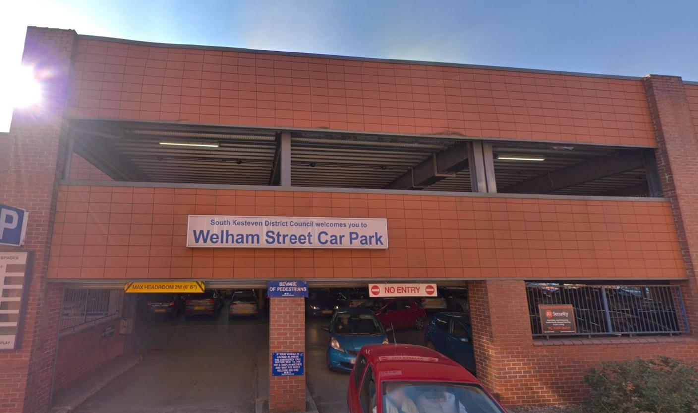 A multi-storey car park entrance and exit made out of orange brick. A red car is exiting, followed by a blue car. A sign reads 'South Kesteven District Council welcomes you to Welham Street Car Park'.