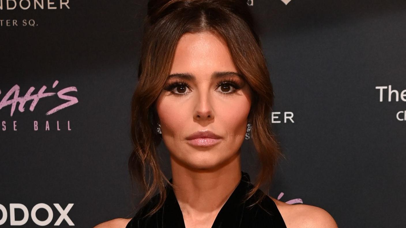 Cheryl Tweedy: the head and shoulders of a woman in front of a black background which is marked with advertising slogans. Her face is framed by brown hair, which is touching her the bottom of her neck. She has brown eyes and is wearing silver earrings and what appears to be a black off-the shoulder dress.  