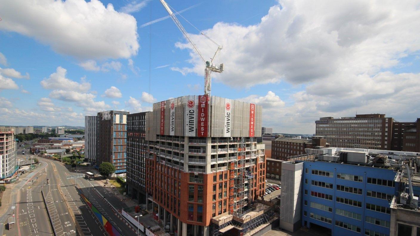 A aerial view of tall building that is under construction. The lower floors are clad in red brick, the middle floors are concrete and still open to the elements, and at the top there are hoardings covering the building with signs that say Winvic. Above the building is a crane