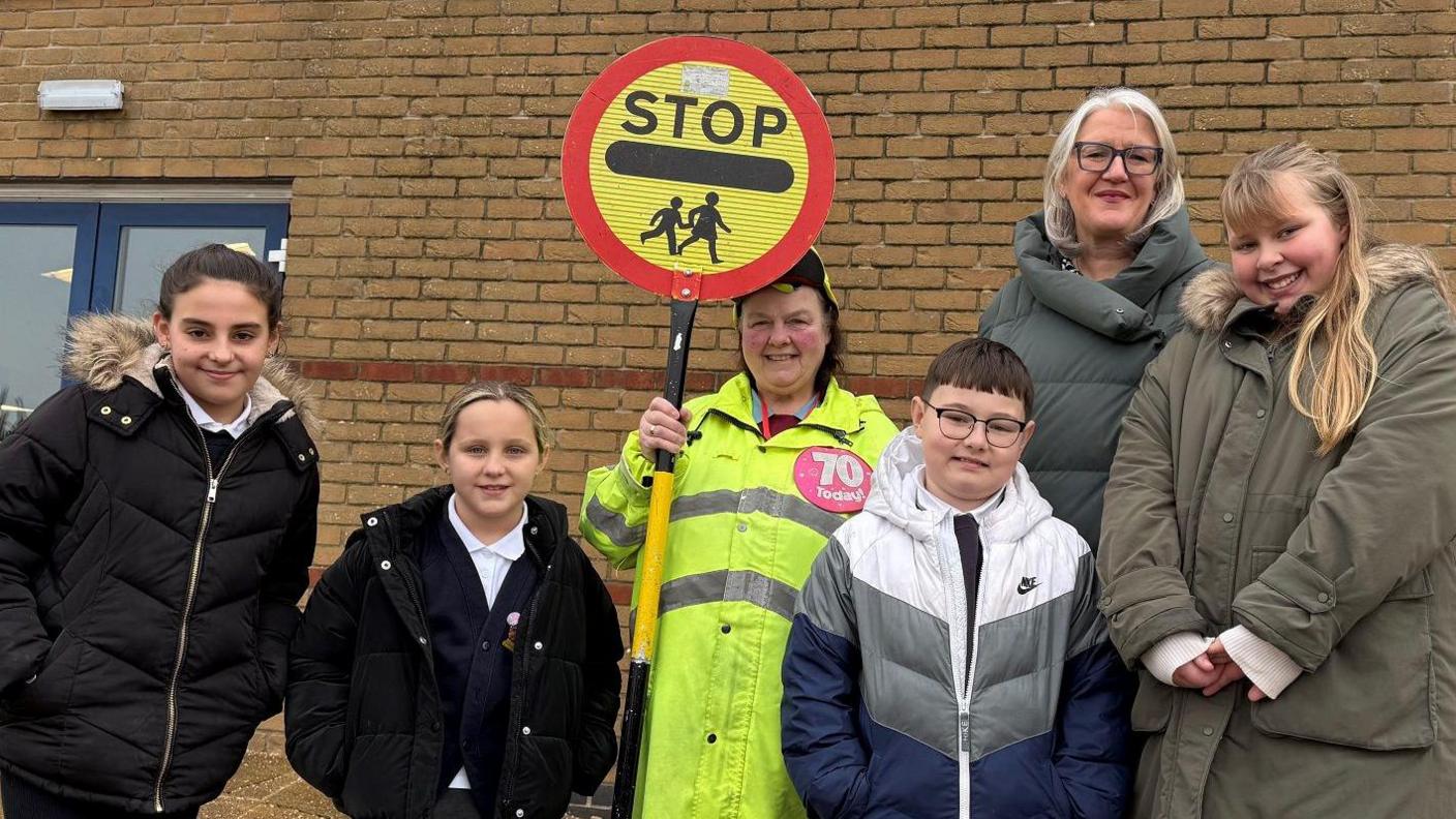 Carol Hawke wearing a red 70 badge in her yellow uniform and with her stop sign. She is surrounded by four school children and the head teacher for a group shot.