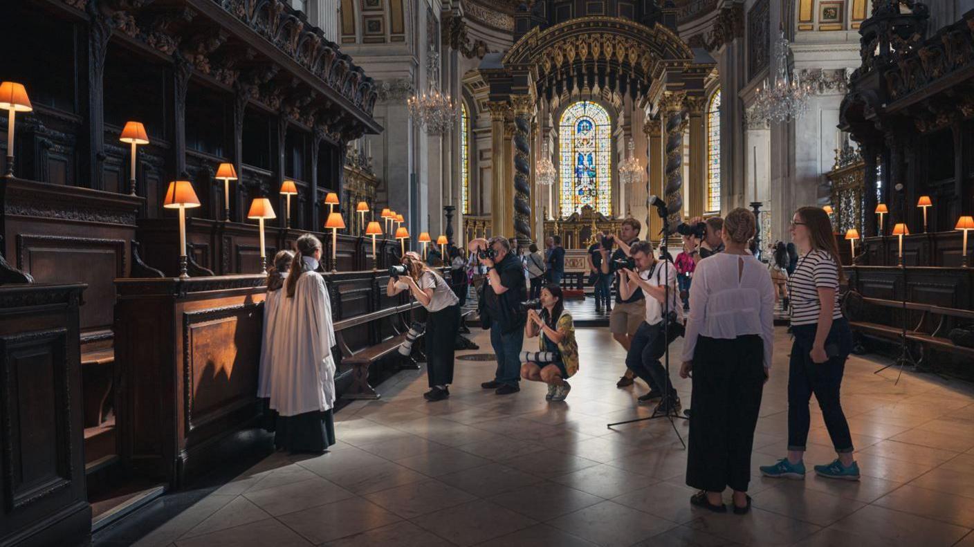 Lila and Lois pose for press photographers in their surplice inside the cathedral. In the background is the large stained glass window and on either side the benches with the reading lamps lit up.