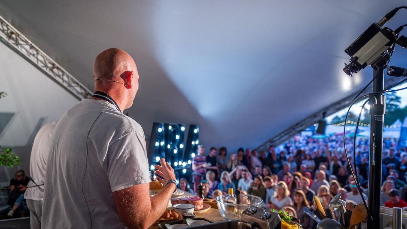 Chef Tom Kerridge in a marquee wearing a white T-shirt standing in front of a crowd watching him demonstrate a recipe