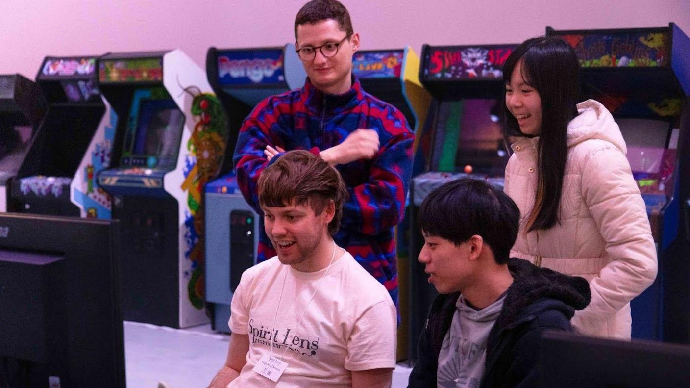 A group of four people are gathered around a gaming console at the WX Exchange. They are all smiling. Retro arcade machines can be seen in the background.