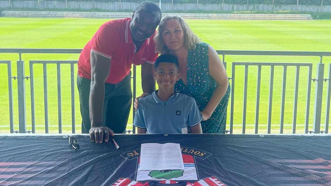 Kayo is sat at a table with a Southampton FC banner on with a contract infront of him. His parents are stood behind him smiling and behind them is a football pitch