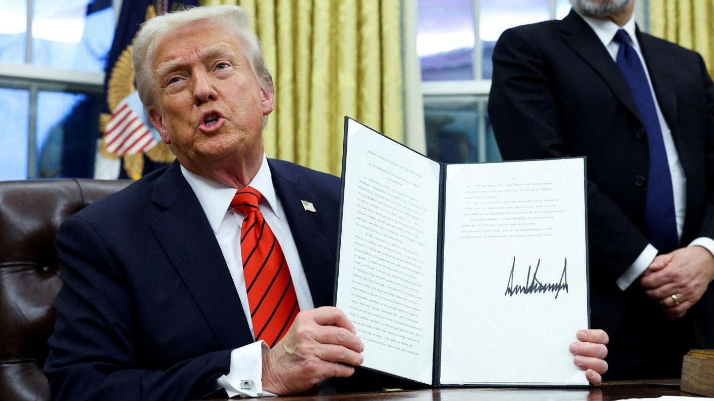 Donald Trump seated behind a desk shows off the text of an executive order.