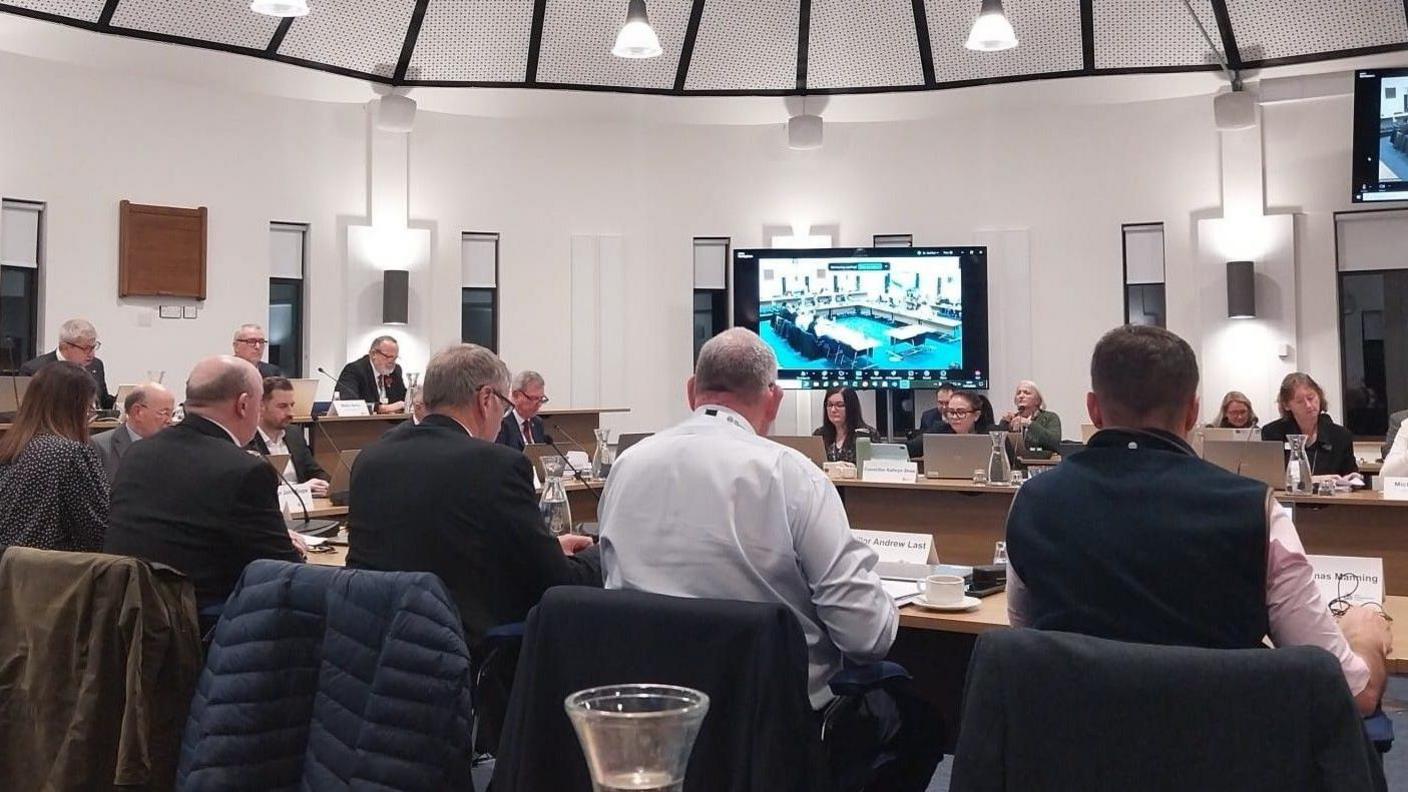 The council chamber at the Forum, with councillors sitting opposite one another. We can see the backs of five people, four men and a women, in the foreground. Futher away are other councillors facing the camera or sideways on. Everyone has a small laptop and a microphone. There are two large TV screens broadcasting the meeting, and the room is modern and well-lit.