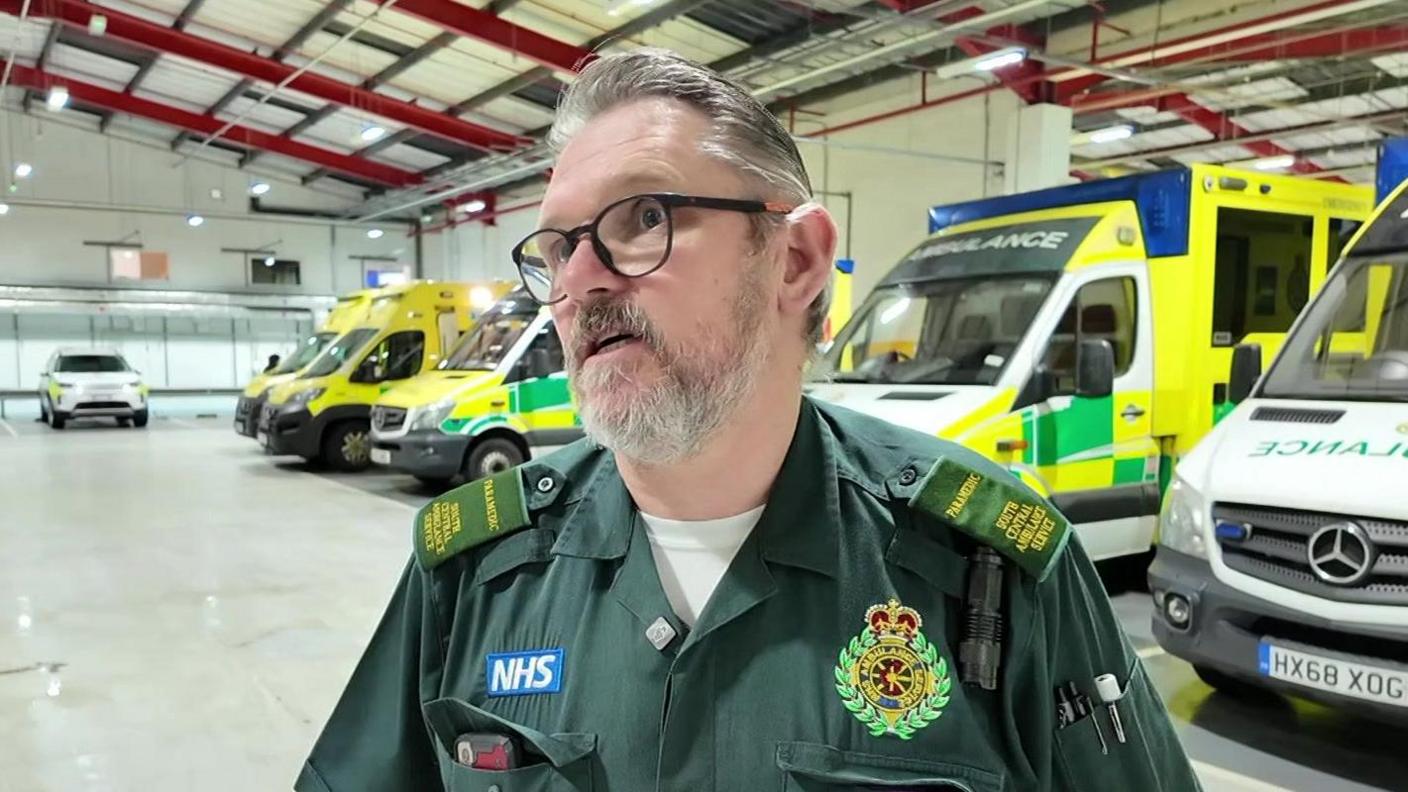 Nigel Young, a man with a grey beard, grey hair and glasses. He is wearing a green NHS paramedic uniform. He is standing in a warehouse and about six ambulances are parked behind him.