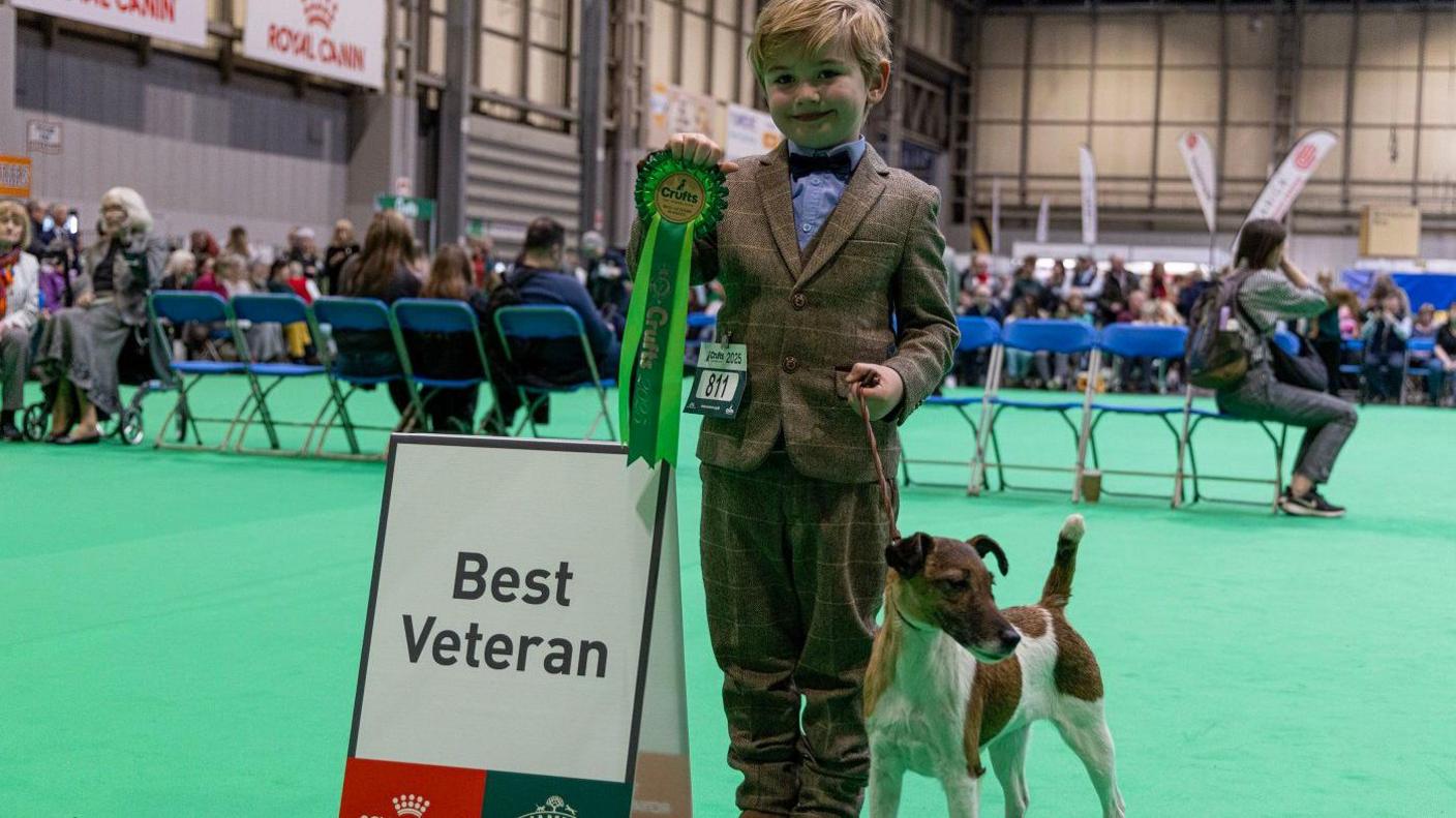 A young boy, Freddie, in a tweed green suit with a blue shirt and dark red bow-tie, stands holding a green rosette. Next to him on a leash is Penny, a dog on all fours with white fur with brown patches. They stand on a light green floor with a row of chairs with some people on behind them. Next to Freddie is a sign with "Best Veteran" written on it.