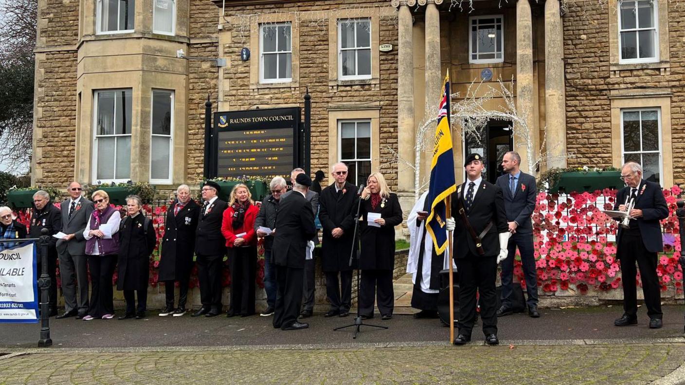 People mostly wearing black standing outside the cream brick Raunds Town Council building. They are lined up in front of the building, which has a display of poppies pinned to a wall. A man holds a flag in front of them.