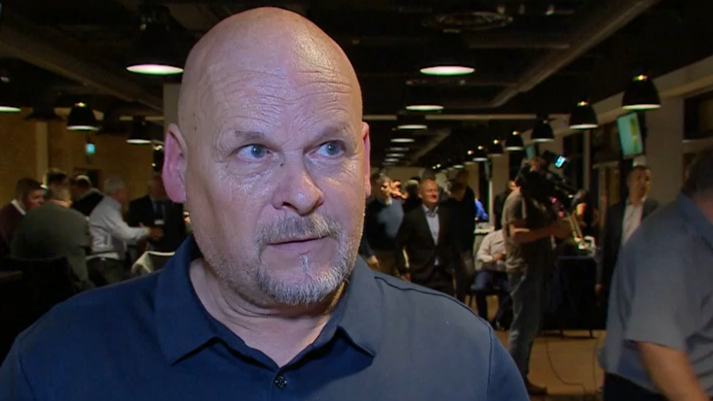 A bald man with a goatee, wearing a dark blue polo-neck shirt, looks to the left of the camera. 