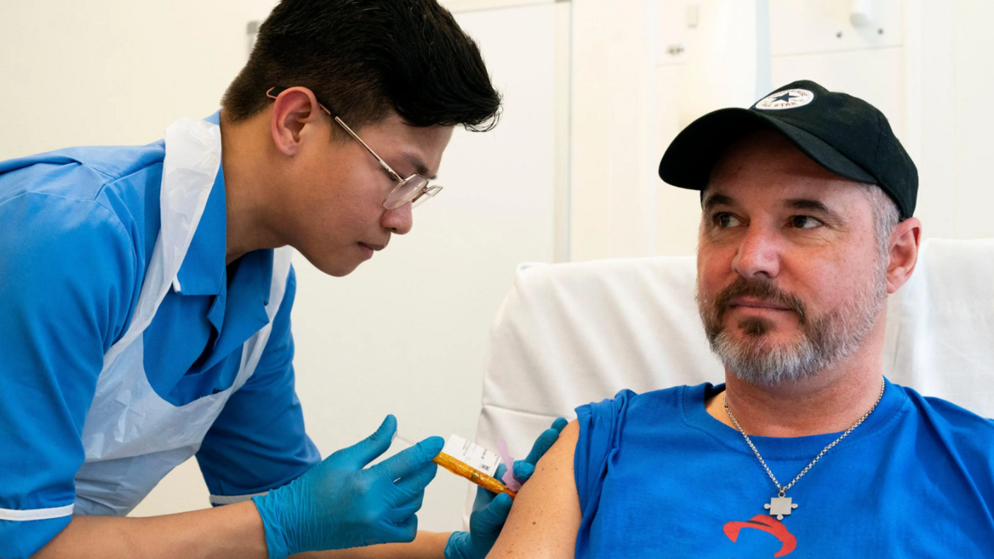 A nurse is on the left of the picture with a needle, inserting it into Steve's arm. Steve is sat on a chair and is wearing a blue top and a black baseball hat. He is wearing a metal chain round his neck with a small jigsaw piece pendant. 