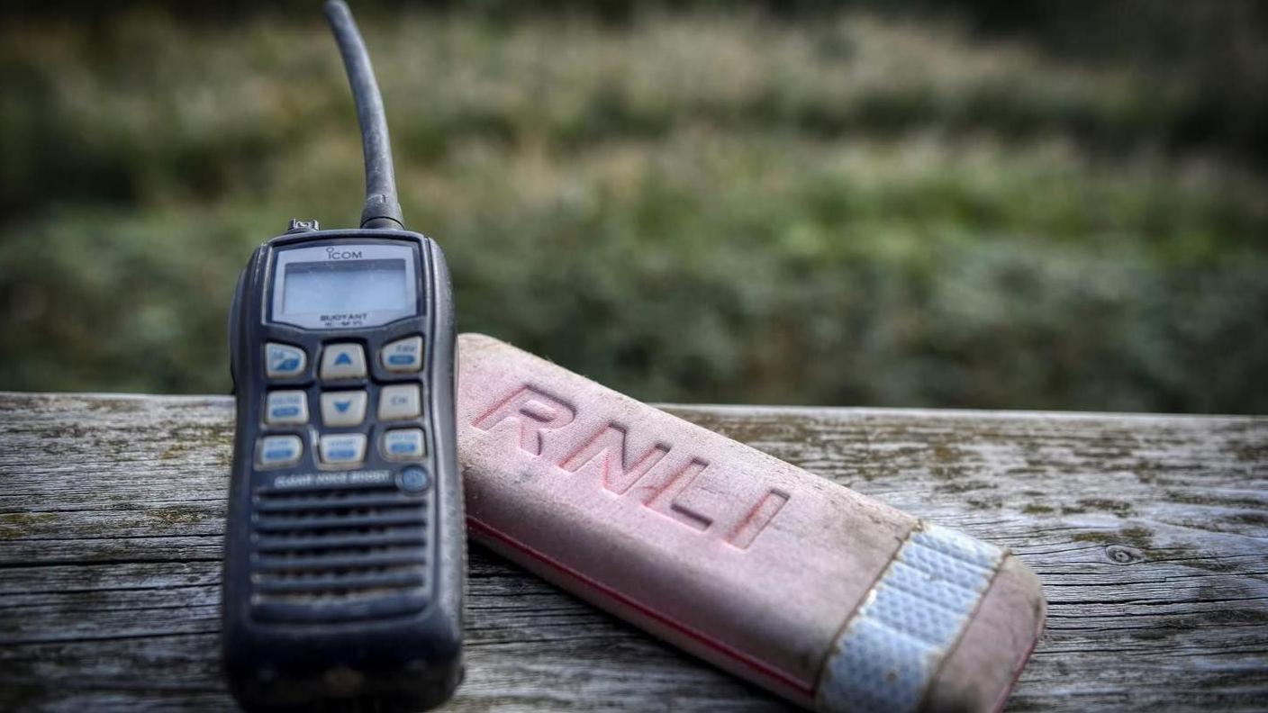 The black walkie-talkie has a short aerial and blue buttons and a faded orange float with the letters RNLI on it. They are laid on a wooden post.