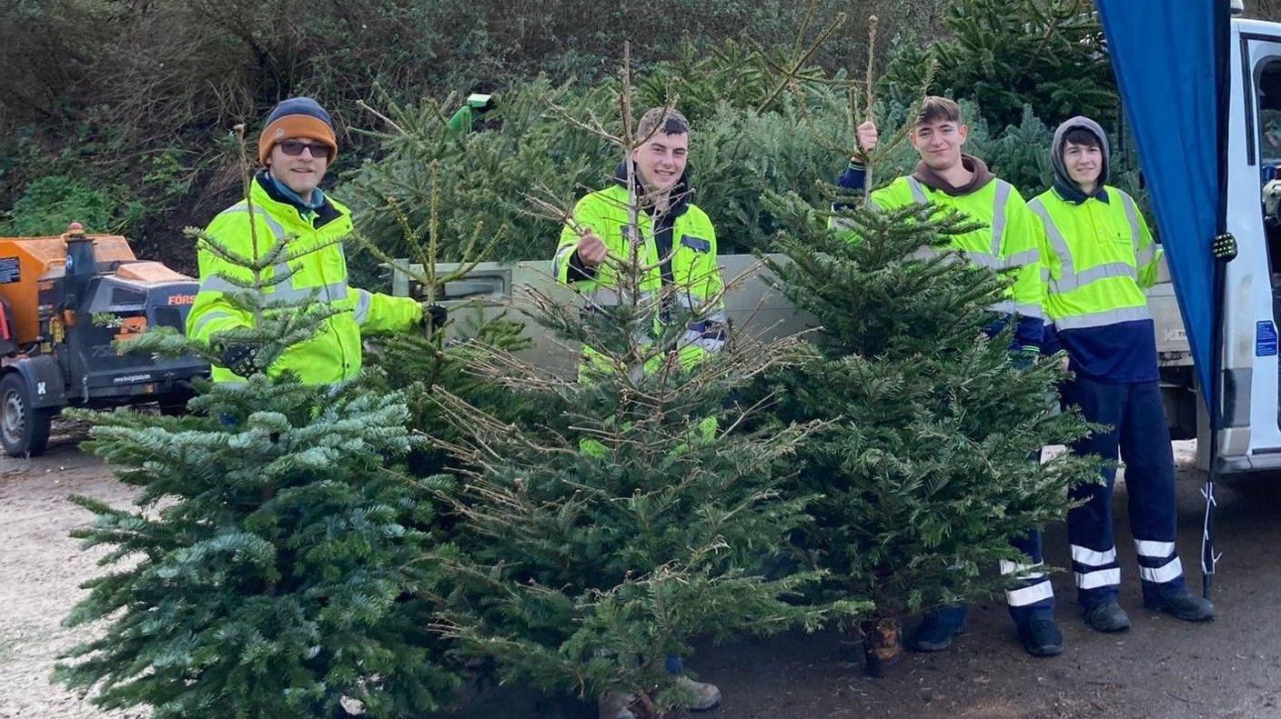 A group of volunteers from St Peter's Hospice in Bristol stand holding short Christmas trees. They are smiling at the camera and are wearing high-vis jackets