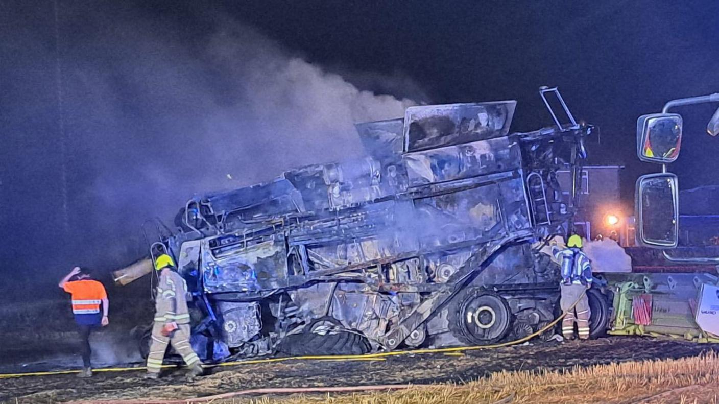 A burnt out combine harvester in a field, firefighters appear to be inspecting the vehicle. There is damage to the ground in the field. It is night time. Smoke is drifting off the vehicle.