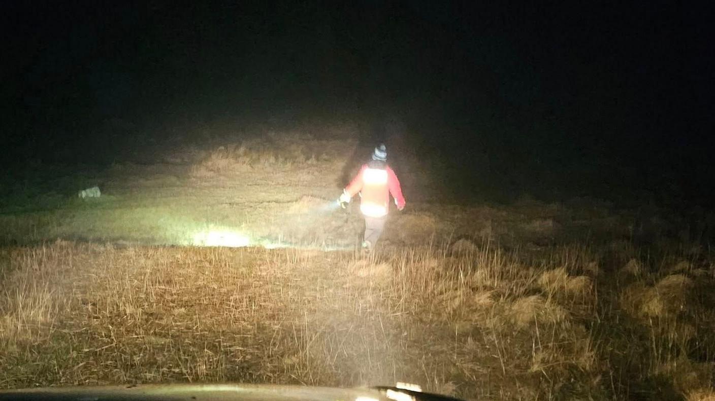 A man is seen from behind walking across moorland with a flashlight illuminating the ground to the side and front of him. He is wearing an orange high-viz jacket 