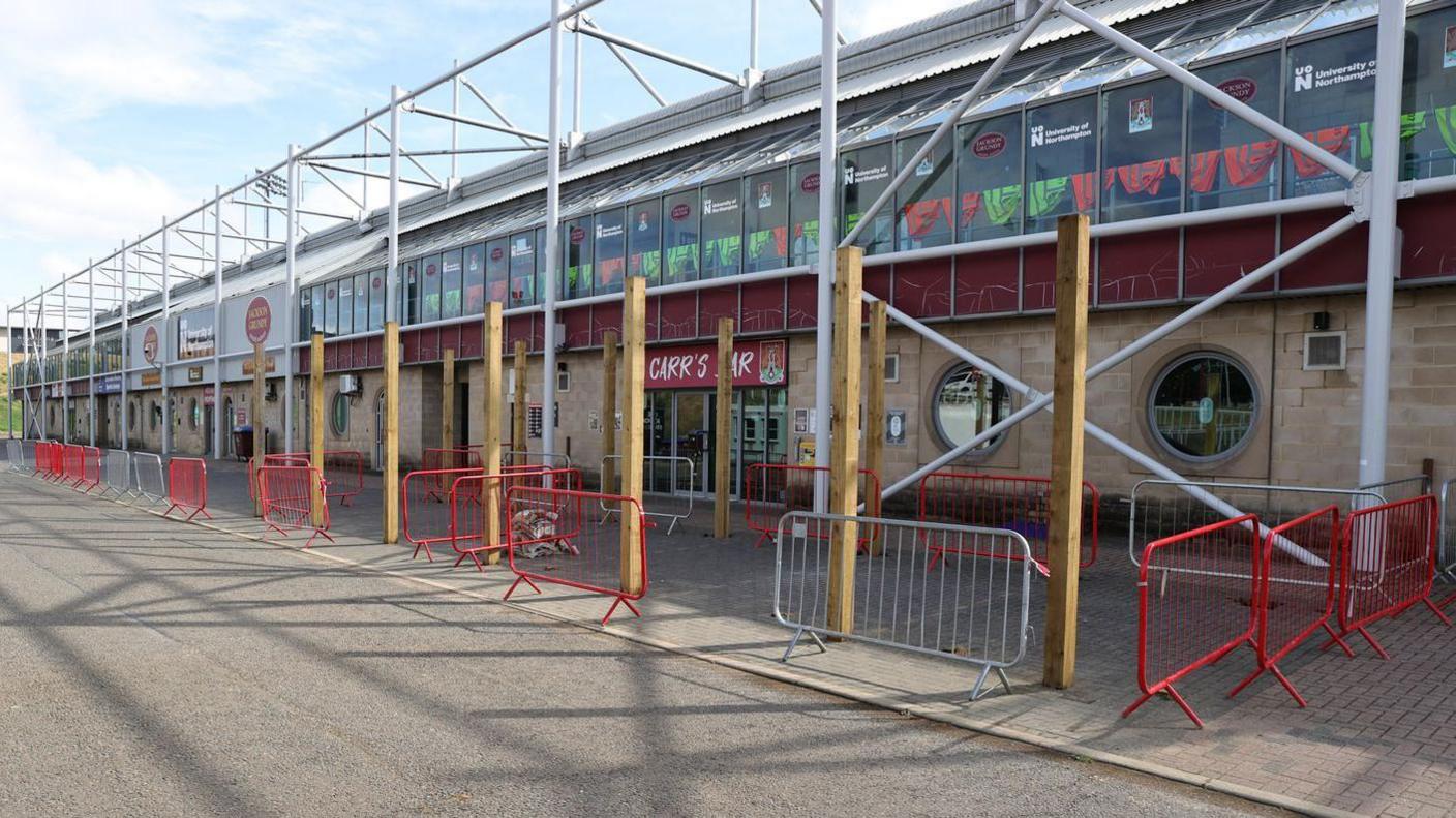 The outside of a football club, with paving and a road in the foreground. There are metal barriers in white and red dotted around the building.