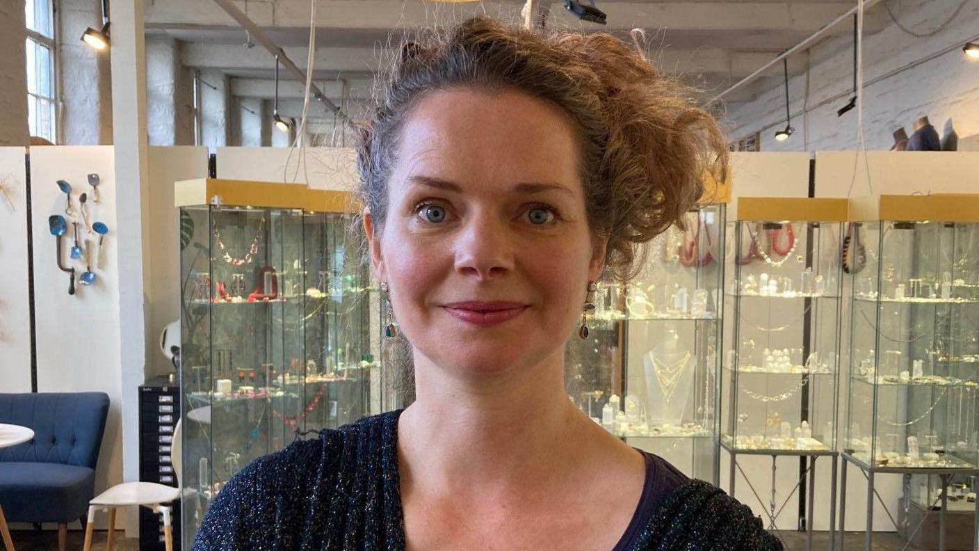 A woman wearing a dark blue cardigan and long hair gathered to one side of her head standing in a jewellery gallery with glass display cabinets.  