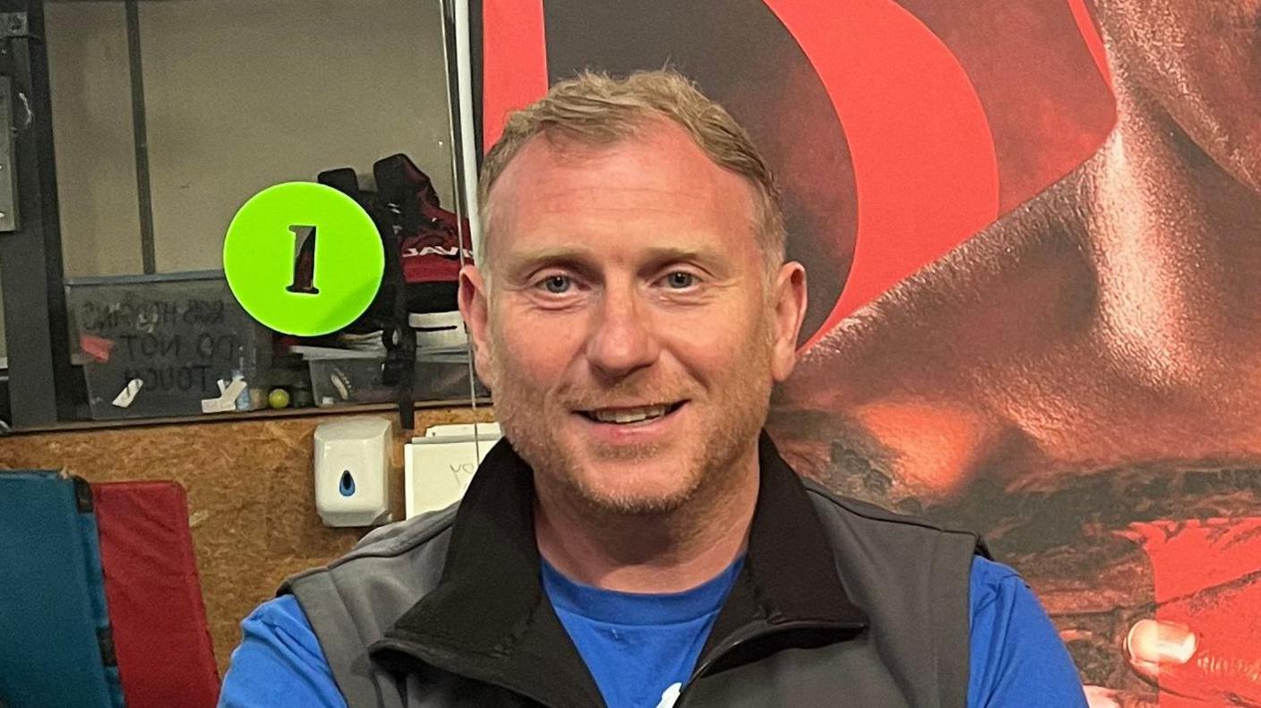 Matt Brennan smiles at the camera. He has short blonde hair and wears a black gilet with a blue T-shirt underneath. He stands in front of a poster board with an image of Fabio Wardley on it, part of which is slightly out of camera.