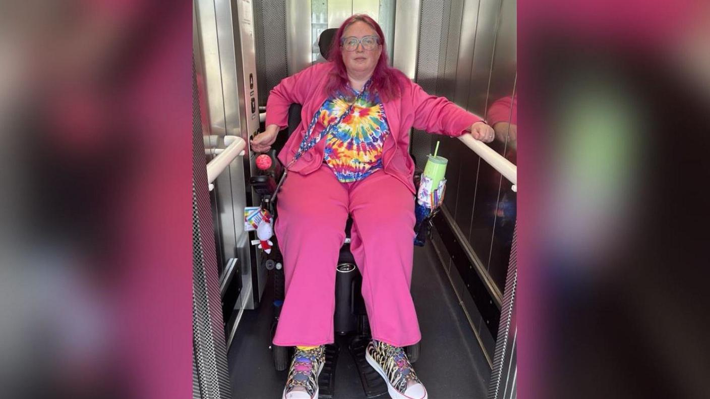 A woman in a bright pink outfit is in a lift in a wheelchair.