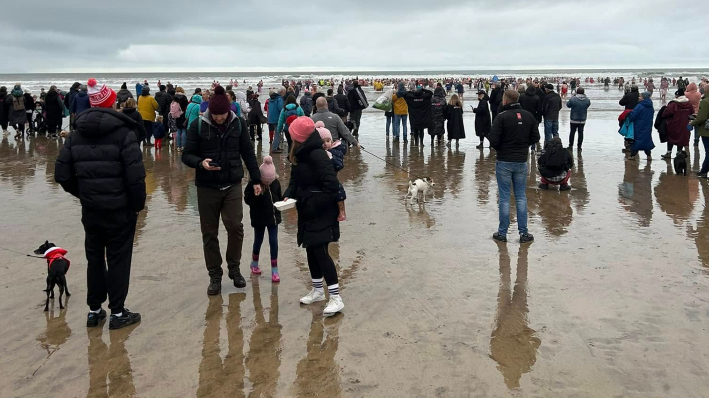 Hundreds of people wearing coats and warm weather outfits standing on a beach as people in the distance head into the sea.