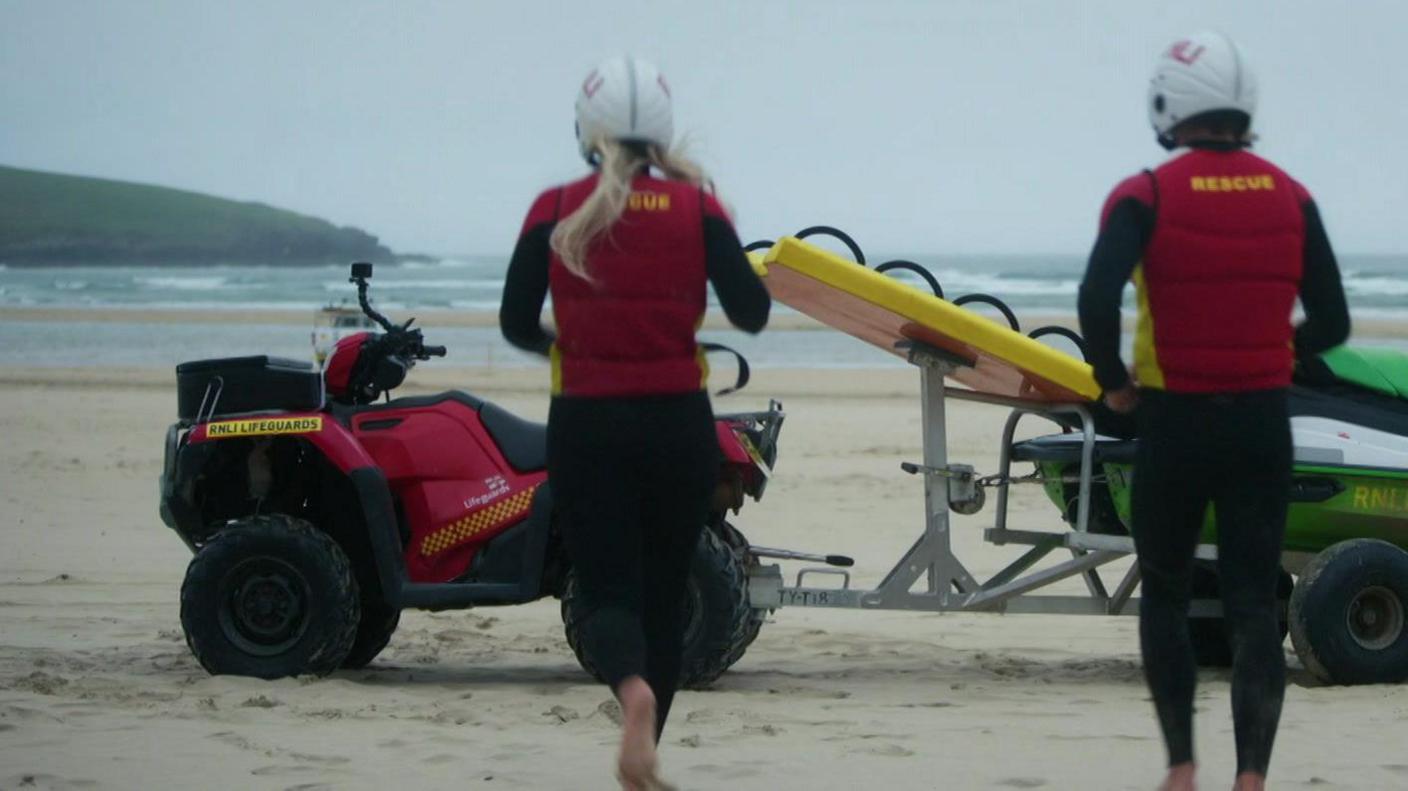 A picture of two people running towards a vehicle parked on the beach. There is two lifeguards pictured both wearing a black and red outfit.