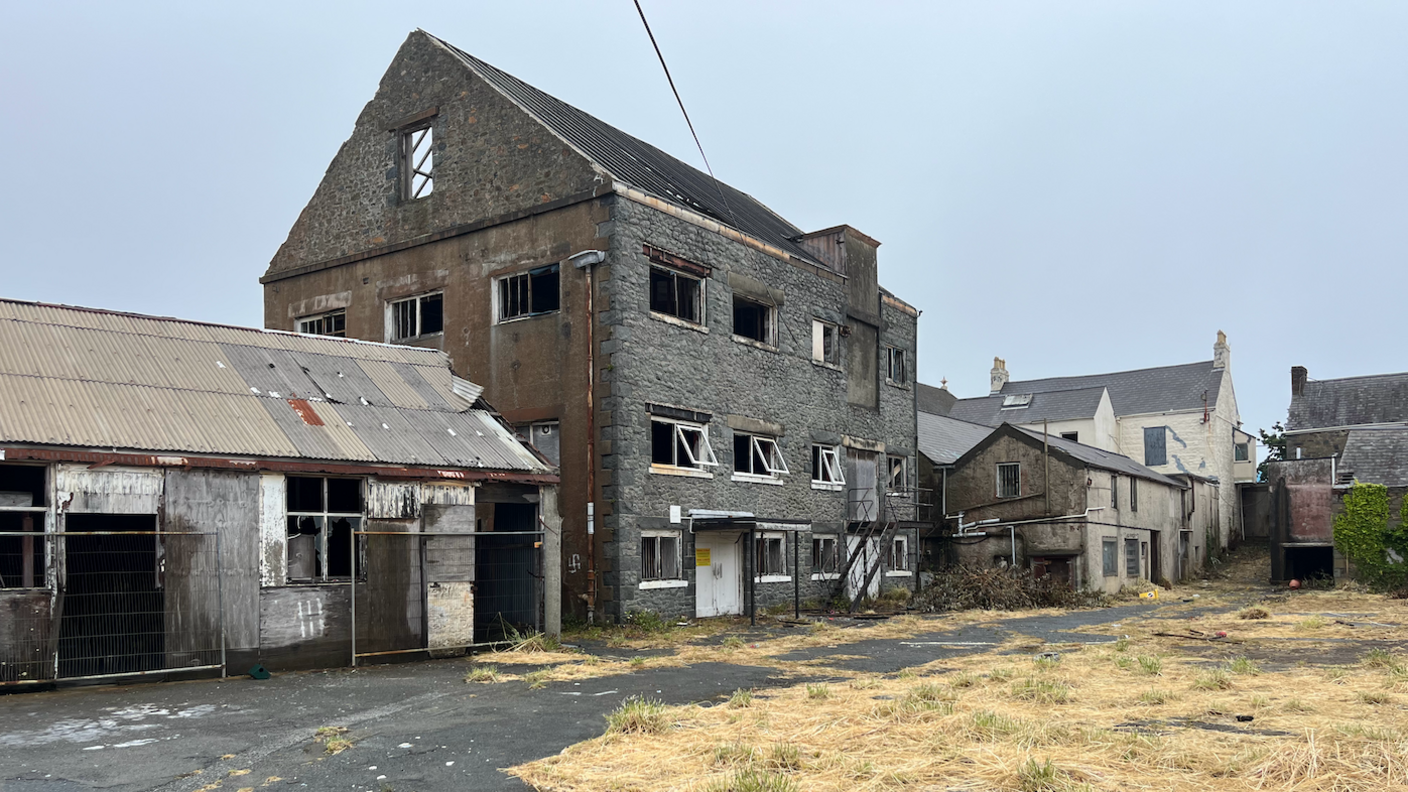 A row of derelict buildings with hay in front of it on the pathway. Grey skies. The grass in front of the building is a light brown.